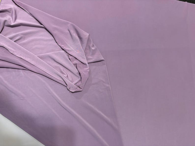 May include: A solid, light purple fabric with a smooth, matte texture. The material is draped on one side, showing soft folds and a consistent, opaque color throughout the surface. The fabric appears lightweight and flexible with a clean finish.