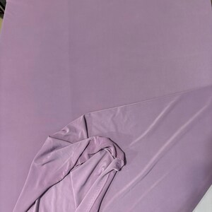 May include: A solid light purple fabric with a smooth, matte texture. The material is draped to show a folded section, revealing the consistent color and opaque quality of the textile. The surface appears uniform with no visible patterns or prints.