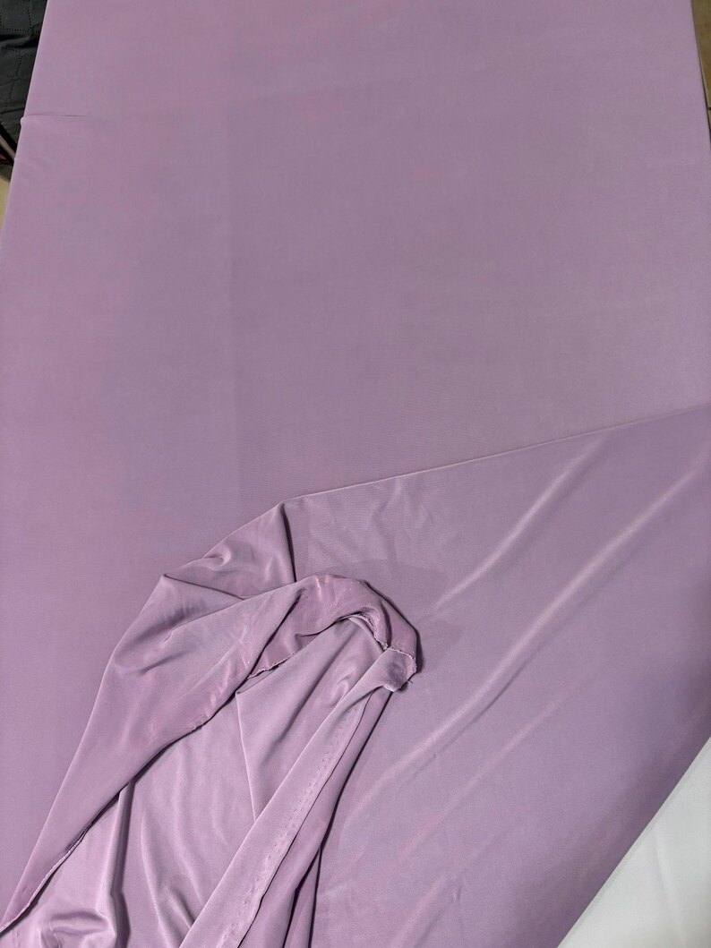 May include: A solid light purple fabric with a smooth, matte texture. The material is draped to show a folded section, revealing the consistent color and opaque quality of the textile. The surface appears uniform with no visible patterns or prints.