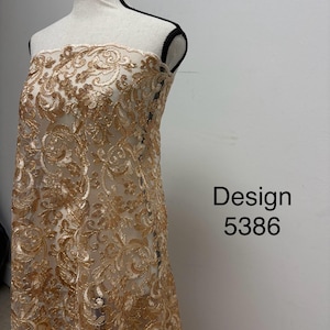 May include: A mannequin draped with gold-colored lace fabric. The lace features an intricate floral design. The text "Design 5386" is visible on the right side of the image. The fabric is semi-sheer, revealing the mannequin underneath.