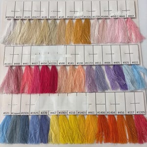 May include: A color chart featuring three rows of thread samples attached to white paper cards. Each sample is labeled with a specific number or color name, displaying a wide spectrum of hues including whites, pinks, yellows, blues, and oranges.