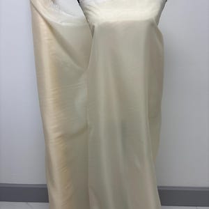 Puede incluir: Un rollo de tela transparente de color marfil se encuentra junto a un maniquí cubierto con el mismo material. La tela tiene un brillo suave y parece ligera. El maniquí es blanco con un ribete de encaje en la parte superior. El fondo es una pared blanca lisa.