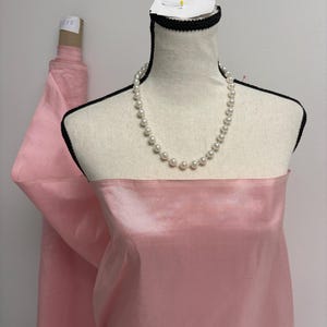 May include: A dress form draped in a smooth, light pink fabric with a subtle sheen. A single strand of white, round faux pearls rests around the neck of the mannequin. The fabric extends from the form, showing a roll of the same pink material.