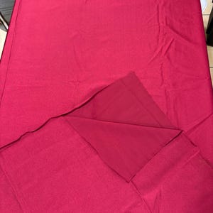 May include: A solid magenta rectangular fabric piece with a smooth, matte texture. The material is folded over at one corner, revealing a consistent deep pink color on both the front and back sides of the textile. The fabric appears flat and even.