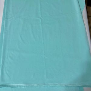 May include: A solid light turquoise fabric with a smooth, matte texture. The material is folded in layers, showing a consistent color throughout. The edges of the fabric are visible at the bottom, displaying a finished, straight-stitched hemline.