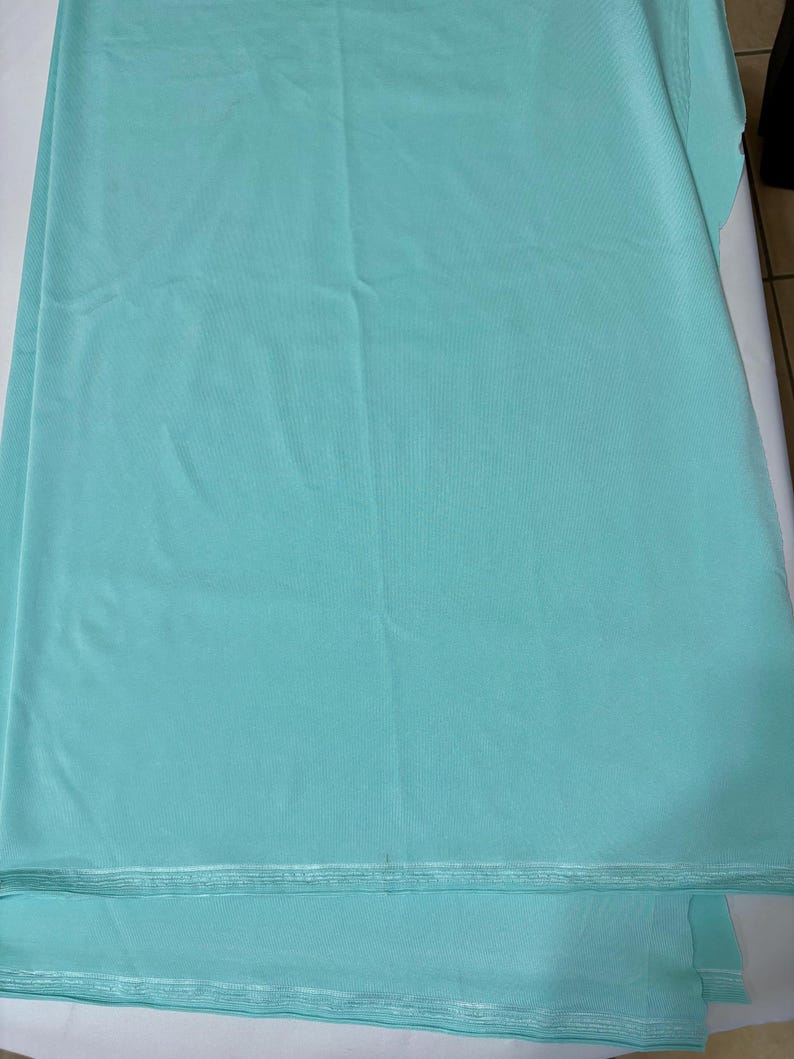 May include: A solid light turquoise fabric with a smooth, matte texture. The material is folded in layers, showing a consistent color throughout. The edges of the fabric are visible at the bottom, displaying a finished, straight-stitched hemline.