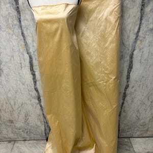 May include: A golden-yellow fabric is draped over a mannequin, showcasing its smooth texture and sheen. A roll of the same fabric is visible to the right. The fabric appears to be a lightweight material, suitable for dresses or other apparel.