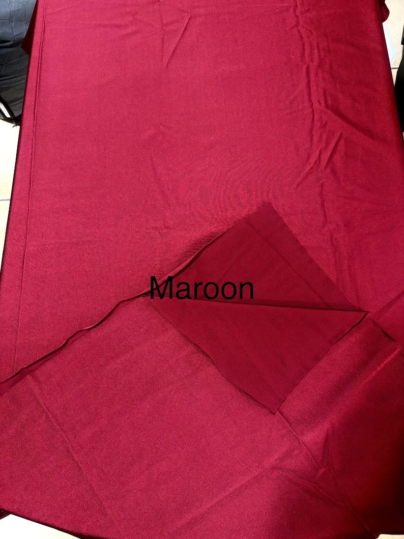 May include: A solid maroon fabric tablecloth with a smooth texture. The fabric is folded over at the corner to reveal the underside, and the word Maroon is printed in black text on the surface of the fabric in the center of the image.