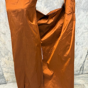 May include: A mannequin displays a burnt orange fabric, possibly silk or satin, with a lustrous sheen. The fabric is draped to suggest a garment, with folds and a flowing appearance. The color is a rich, warm tone, and the fabric appears to be of high quality.