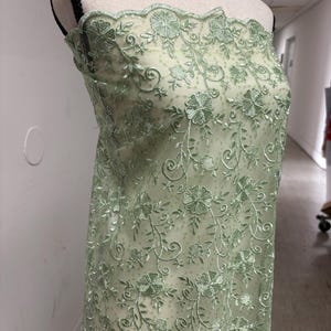Op de afbeelding: Een lichtgroene bloemen geborduurde stof gedrapeerd over een mannequin. De delicate borduursels hebben een herhalend patroon van bloemen en wijnranken. De stof heeft een geschulpte rand en een transparante uitstraling, wat suggereert dat deze voor kleding of decoratieve doeleinden gebruikt kan worden.