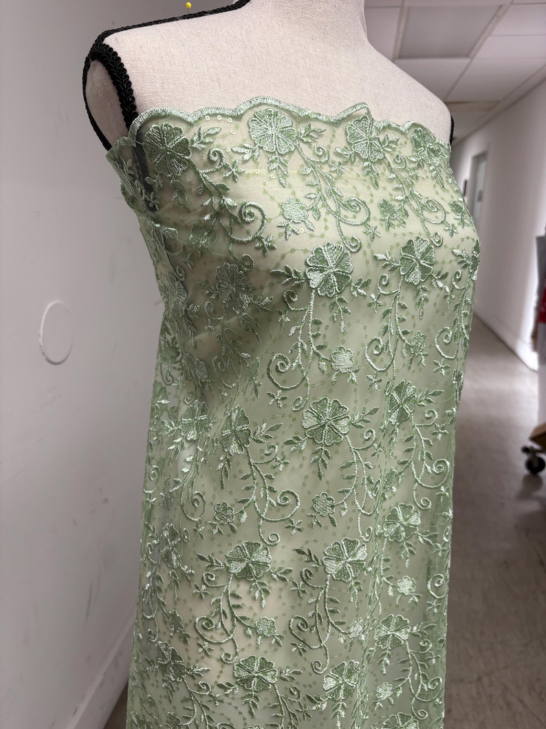 Op de afbeelding: Een lichtgroene bloemen geborduurde stof gedrapeerd over een mannequin. De delicate borduursels hebben een herhalend patroon van bloemen en wijnranken. De stof heeft een geschulpte rand en een transparante uitstraling, wat suggereert dat deze voor kleding of decoratieve doeleinden gebruikt kan worden.