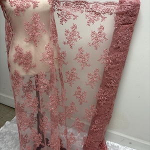K&ouml;nnte beinhalten: Staubrosa Spitzenstoff mit floraler Stickerei. Der Stoff ist &uuml;ber eine Schaufensterpuppe drapiert und aufgerollt, wodurch die komplizierten Details des Designs zur Geltung kommen. Die Spitze hat einen gewellten Rand und ein zartes, offenes Gewebe.