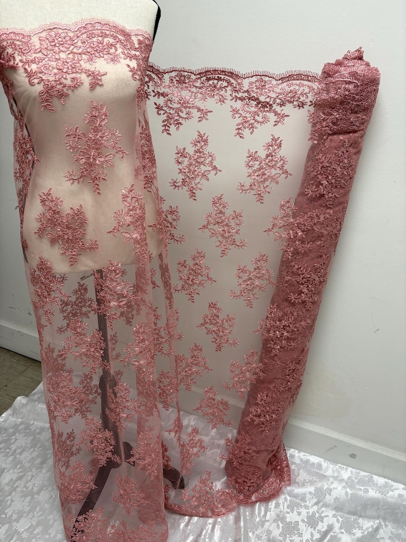 K&ouml;nnte beinhalten: Staubrosa Spitzenstoff mit floraler Stickerei. Der Stoff ist &uuml;ber eine Schaufensterpuppe drapiert und aufgerollt, wodurch die komplizierten Details des Designs zur Geltung kommen. Die Spitze hat einen gewellten Rand und ein zartes, offenes Gewebe.
