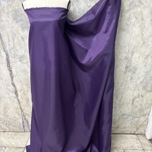 May include: A length of vivid purple fabric draped over a mannequin. The material has a smooth, glossy texture and a raw edge. The mannequin is white with a black trim.
