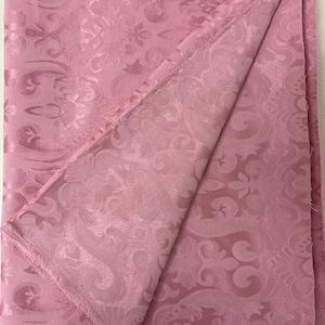 May include: A folded pink fabric with a detailed floral and scroll pattern. The fabric has a subtle sheen and a fringed edge. The design is visible on both sides of the folded material, suggesting a double-sided textile.