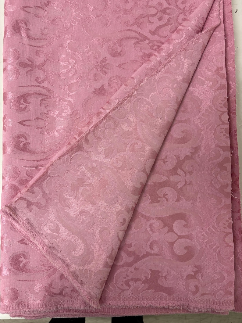 May include: A folded pink fabric with a detailed floral and scroll pattern. The fabric has a subtle sheen and a fringed edge. The design is visible on both sides of the folded material, suggesting a double-sided textile.