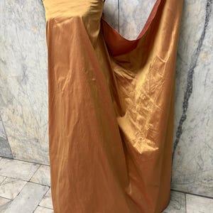 May include: A draped, golden-orange fabric is displayed on a mannequin. The fabric has a lustrous sheen and appears to be silk or a similar material. The fabric is draped to create a strapless dress or gown.