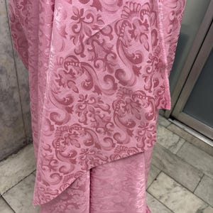 May include: A close-up of pink fabric with a repeating floral damask pattern. The fabric has a slight sheen, and the pattern is a darker shade of pink, creating a subtle contrast. The fabric is draped, showing folds and texture.