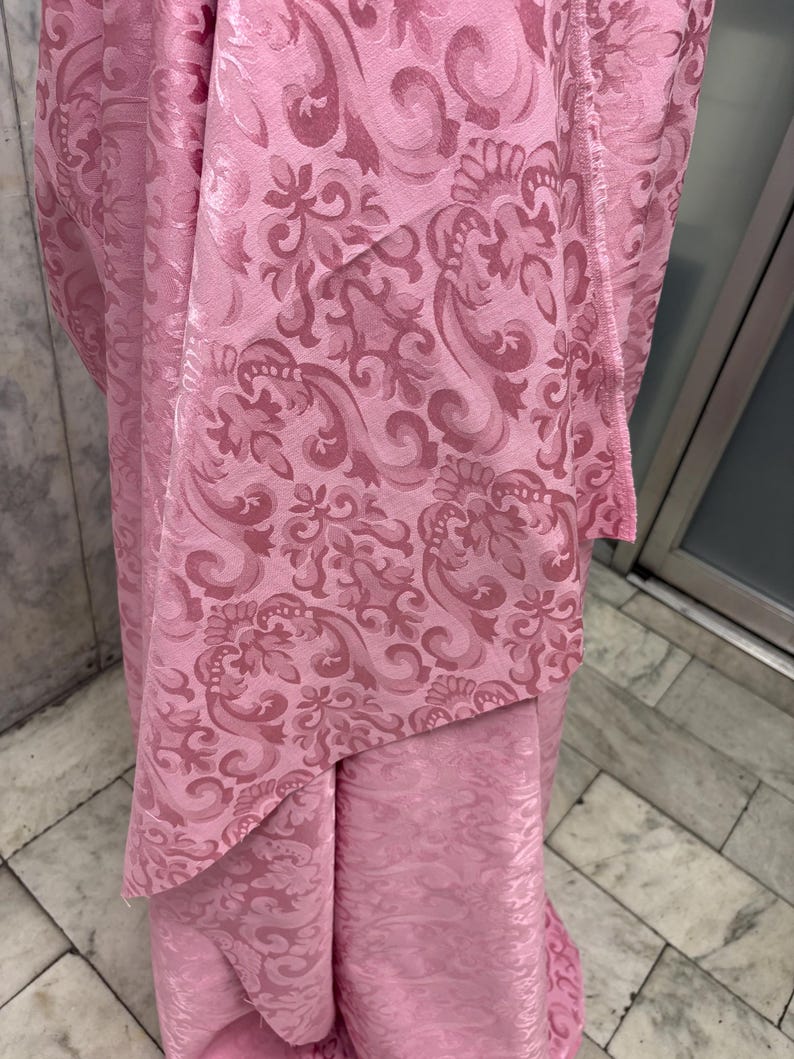 May include: A close-up of pink fabric with a repeating floral damask pattern. The fabric has a slight sheen, and the pattern is a darker shade of pink, creating a subtle contrast. The fabric is draped, showing folds and texture.