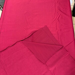 May include: A solid deep magenta fabric tablecloth with a smooth, matte texture. The rectangular textile is draped flat, with one corner folded back to reveal the underside of the same consistent, vibrant pinkish-red color and material.