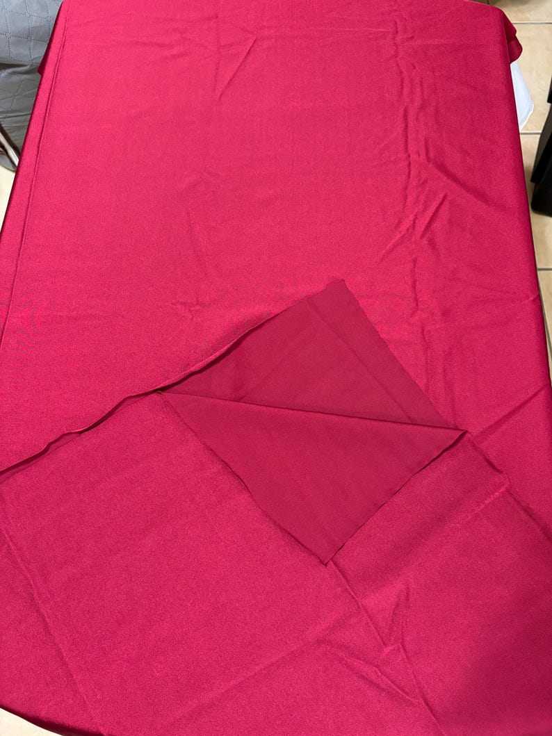 May include: A solid deep magenta fabric tablecloth with a smooth, matte texture. The rectangular textile is draped flat, with one corner folded back to reveal the underside of the same consistent, vibrant pinkish-red color and material.