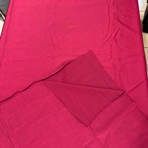 May include: A solid deep magenta fabric tablecloth with a smooth, matte texture. The rectangular textile is draped flat, with one corner folded back to reveal the underside of the same consistent, vibrant pinkish-red color and material.