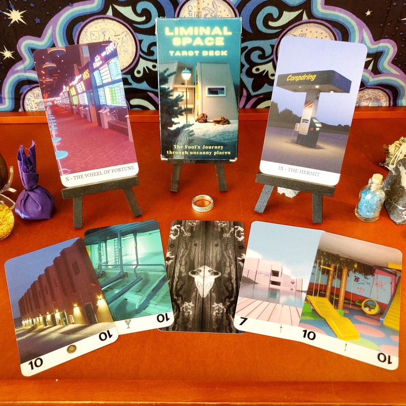 Five Liminal Space tarot cards rest flat on a wooden platform with two set on stands behind them and the box on a stand in the middle. The cards show liminal spaces, such as the suburbs, playrooms, and poolrooms.