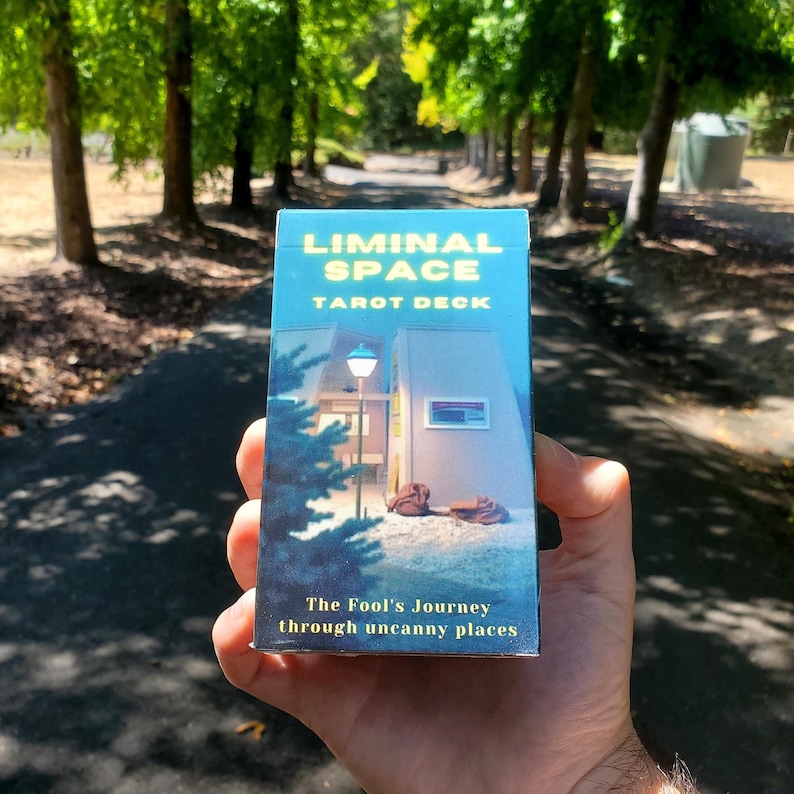 The front of the Liminal Space Tarot Deck held up against a background of a road lined with green leafy trees.