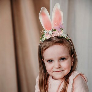 Puede incluir: Una niña pequeña con un vestido rosa, una diadema floral y orejas de conejo. La diadema tiene flores rosas, hojas verdes y una mariposa.