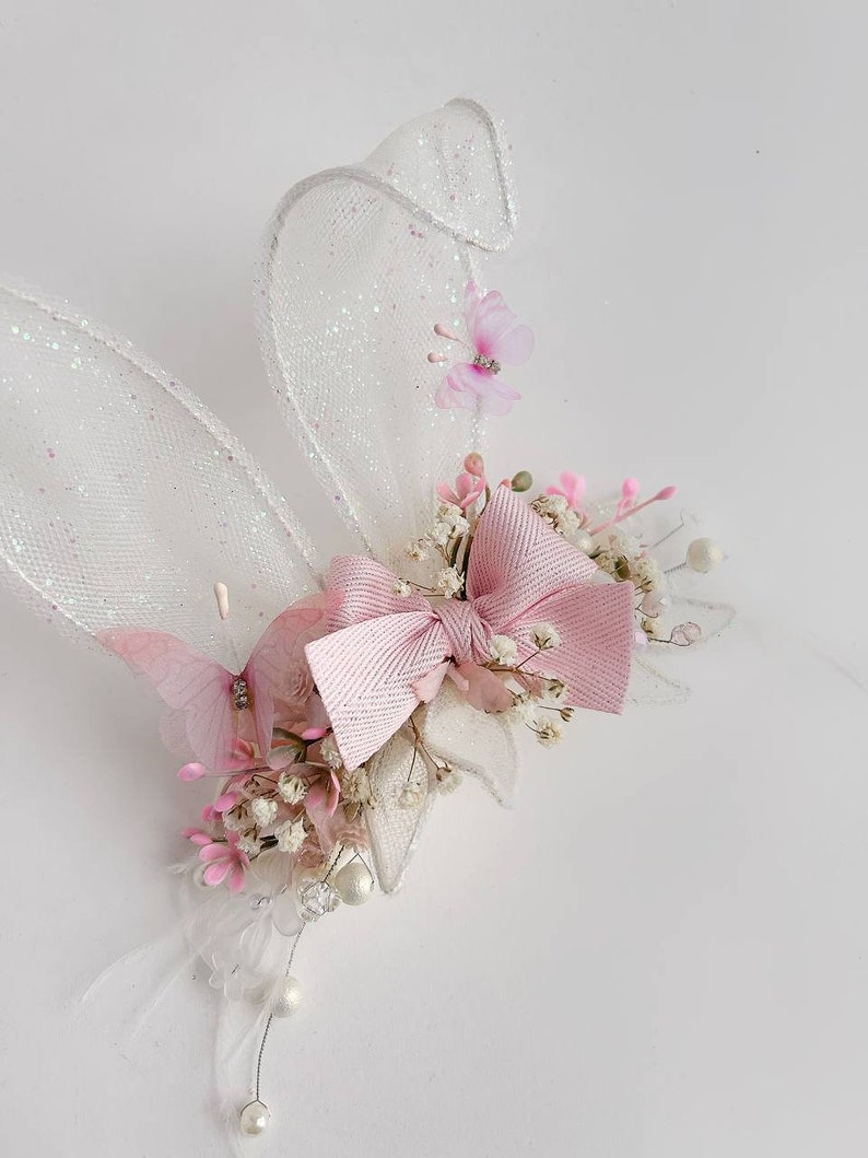 K&ouml;nnte beinhalten: Ein rosa-wei&szlig;es Hasenohr-Haarband mit einer rosa Schleife, wei&szlig;em T&uuml;ll und rosa Blumen.