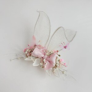K&ouml;nnte beinhalten: Eine zarte Haarklammer mit einer rosa Schleife, wei&szlig;en Bl&uuml;ten und durchsichtigen wei&szlig;en Fl&uuml;geln mit Glitzerakzenten.
