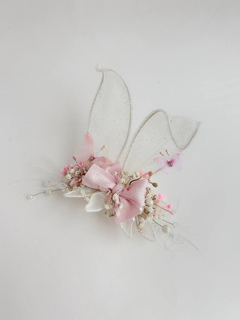 K&ouml;nnte beinhalten: Eine zarte Haarklammer mit einer rosa Schleife, wei&szlig;en Bl&uuml;ten und durchsichtigen wei&szlig;en Fl&uuml;geln mit Glitzerakzenten.