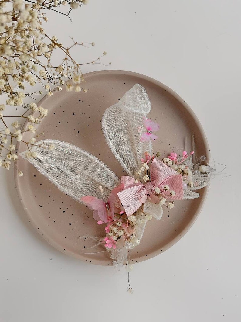 K&ouml;nnte beinhalten: Eine zarte Haarklammer mit einer rosa Schleife, wei&szlig;en T&uuml;llfl&uuml;geln und getrockneten Blumen. Die Klammer ist perfekt, um jeder Frisur einen Hauch von Verspieltheit zu verleihen.