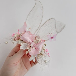 K&ouml;nnte beinhalten: Eine zarte rosa und wei&szlig;e Blumenhaarspange mit einer rosa Schleife und Schmetterlingsakzenten. Die Spange besteht aus einem durchsichtigen wei&szlig;en Stoff mit glitzernder Oberfl&auml;che, der an Schmetterlingsfl&uuml;gel erinnert.
