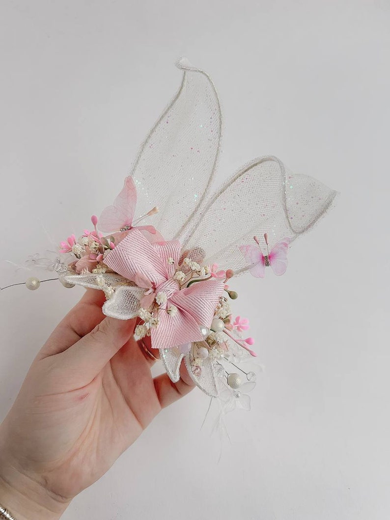 K&ouml;nnte beinhalten: Eine zarte rosa und wei&szlig;e Blumenhaarspange mit einer rosa Schleife und Schmetterlingsakzenten. Die Spange besteht aus einem durchsichtigen wei&szlig;en Stoff mit glitzernder Oberfl&auml;che, der an Schmetterlingsfl&uuml;gel erinnert.