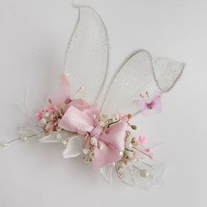K&ouml;nnte beinhalten: Eine rosafarbene Schleife mit wei&szlig;en Bl&uuml;ten und zwei glitzernden wei&szlig;en Fl&uuml;geln mit rosa Schmetterlingen.