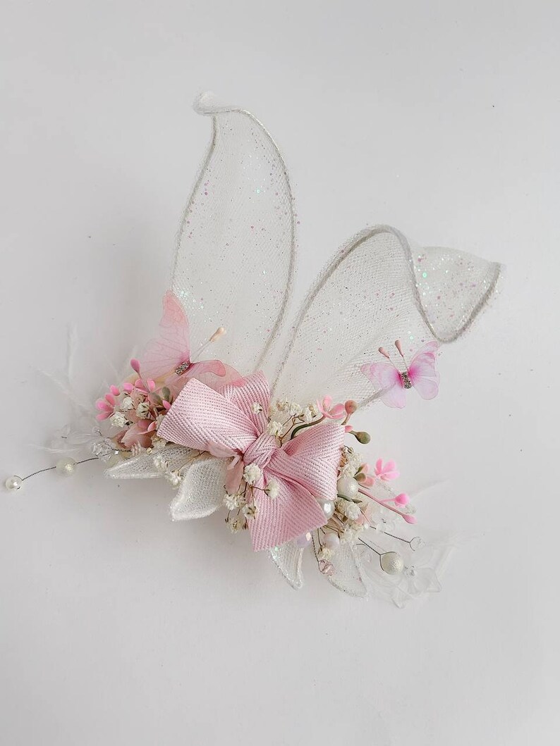 K&ouml;nnte beinhalten: Eine rosafarbene Schleife mit wei&szlig;en Bl&uuml;ten und zwei glitzernden wei&szlig;en Fl&uuml;geln mit rosa Schmetterlingen.