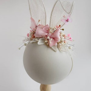 K&ouml;nnte beinhalten: Ein rosa-wei&szlig;es Stirnband mit Hasenohren, einer rosa Schleife und wei&szlig;en Blumen. Das Stirnband befindet sich auf einem wei&szlig;en Mannequin-Kopf.
