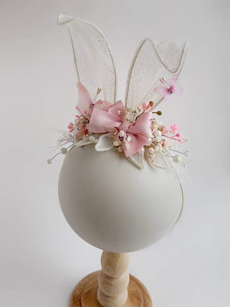 K&ouml;nnte beinhalten: Ein rosa-wei&szlig;es Stirnband mit Hasenohren, einer rosa Schleife und wei&szlig;en Blumen. Das Stirnband befindet sich auf einem wei&szlig;en Mannequin-Kopf.