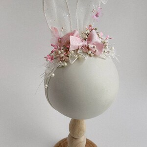 K&ouml;nnte beinhalten: Ein wei&szlig;es Stirnband mit rosa Hasenohren, einer rosa Schleife und wei&szlig;en und rosa Blumen. Das Stirnband befindet sich auf einem Holzst&auml;nder.