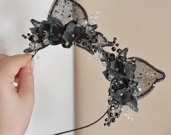 May include: A black headband with decorative cat ears. The ears are made of black lace with a dotted pattern and are adorned with black flowers, beads, and crystals. The headband is held by a hand.