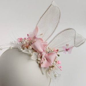 K&ouml;nnte beinhalten: Ein wei&szlig;es Stirnband mit rosa und wei&szlig;en Blumen, einer rosa Schleife und wei&szlig;en T&uuml;ll-Hasenohren. Das Stirnband ist mit kleinen wei&szlig;en Perlen verziert.