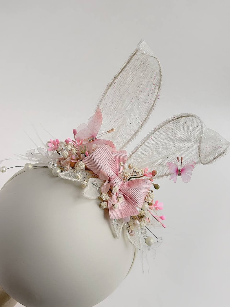K&ouml;nnte beinhalten: Ein wei&szlig;es Stirnband mit rosa und wei&szlig;en Blumen, einer rosa Schleife und wei&szlig;en T&uuml;ll-Hasenohren. Das Stirnband ist mit kleinen wei&szlig;en Perlen verziert.