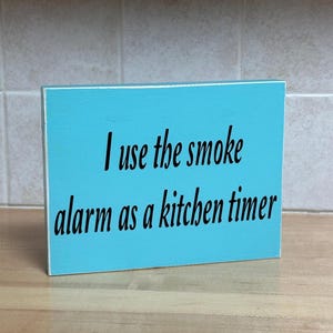 May include: A rectangular, turquoise wooden sign with white edges. The black text reads, "I use the smoke alarm as a kitchen timer." The sign is displayed on a light-colored wooden surface.