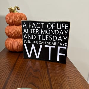 May include: A black and white wooden sign with the text "A FACT OF LIFE AFTER MONDAY AND TUESDAY EVEN THE CALENDAR SAYS WTF". A stack of three orange pumpkins with gold stems is to the left of the sign.