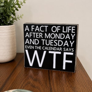 May include: A rectangular black wooden sign with white text that reads: A FACT OF LIFE AFTER MONDAY AND TUESDAY EVEN THE CALENDAR SAYS WTF. The sign has a distressed finish along the edges and is positioned upright on a flat surface.
