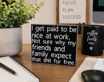 May include: A rectangular black wooden sign with white text that reads: I get paid to be nice at work. Not sure why my friends and family expect that shit for free. The sign has a smooth finish and clean edges, standing upright on a flat surface.