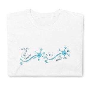 May include: White t-shirt featuring a light blue illustration of neurons connected by a wire. The text "Neurons that fire together wire together" is printed in a matching blue color. The shirt is folded.