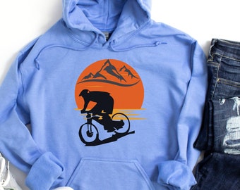 May include: A light blue hoodie with a black graphic of a mountain biker silhouetted against an orange sunset and mountains. The hoodie is paired with blue jeans and gray sneakers.