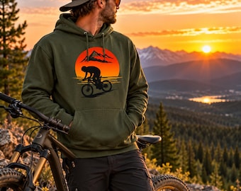 May include: An olive green hooded sweatshirt featuring a graphic print of a cyclist riding a mountain bike in front of a large orange sun and black mountain silhouette. The hoodie includes a front kangaroo pocket and matching drawstrings.
